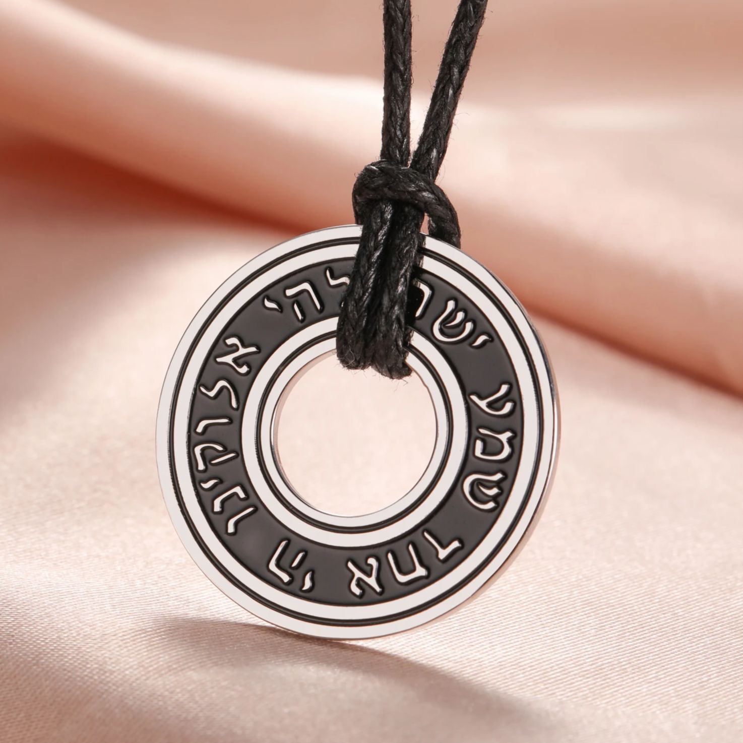 Pendentif rond en acier inoxydable avec inscription hébraïque, monté sur cordon noir réglable.