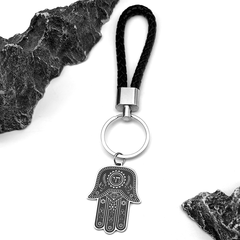 Porte-clés Hamsa en acier inoxydable avec cordon noir tressé et détails hébraïques.